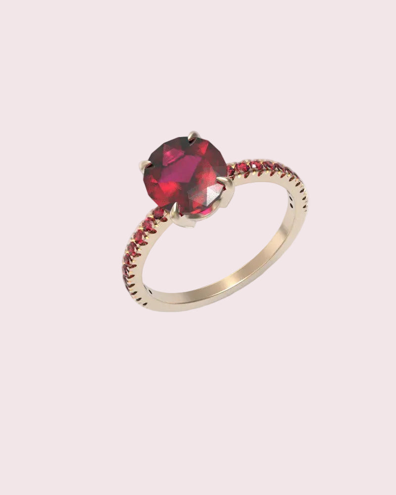 A rose gold ring featuring a large, round-cut deep red gemstone in a prong setting, with a shank accented by small red stones, shown on a clean white background.
